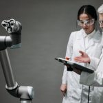 Pesquisadores de jaleco e óculos de proteção analisam um braço robótico em um laboratório.