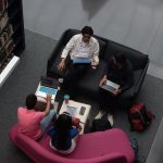 Grupo diverso de adultos colabora em laptops na área de convivência de uma biblioteca.