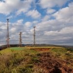 Torres de telecomunicações instaladas no alto de uma colina coberta por vegetação, sob céu nublado.
