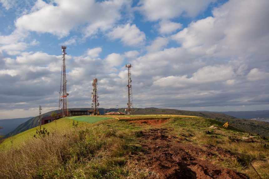 Torres de telecomunicações instaladas no alto de uma colina coberta por vegetação, sob céu nublado.