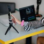 Mulher ajusta um smartphone em um tripé ao lado de um laptop em um ambiente de produção e edição de vídeos.