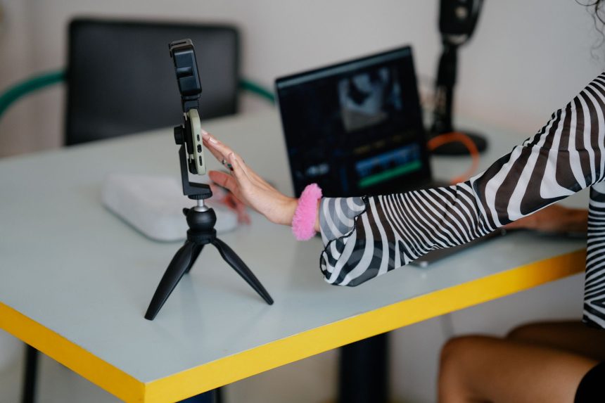 Mulher ajusta um smartphone em um tripé ao lado de um laptop em um ambiente de produção e edição de vídeos.